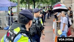 大批穿上防刺戰術背後的軍裝警員在西九龍裁判法院外，列隊監視排隊輪候法庭旁聽席的市民，準備進入法院大樓。(美國之音)