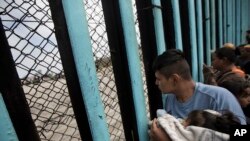 一个中美洲移民车队的人抱着孩子向边界墙对面张望（2018年4月29日）