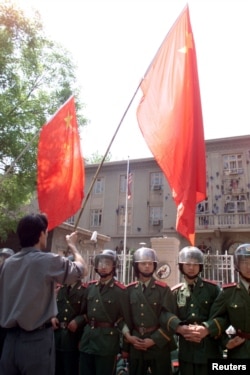 1999年5月10日，中国示威者在美国驻华使馆外面挥舞国旗。中国武警列队保护使馆。使馆墙壁被抗议美国轰炸中国驻南斯拉夫使馆的人用砖石砸得伤痕累累
